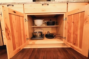 Un mueble de cocina de madera con puertas abiertas, mostrando un estante con utensilios de cocina y una olla.
