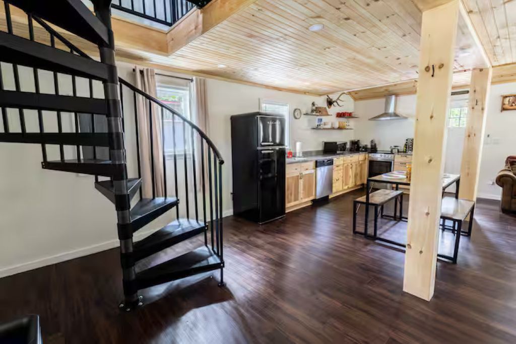 A modern kitchen with dark wood flooring, a black spiral staircase, and a dining area with wooden tables.