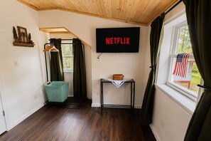 A room with a wooden ceiling, a wall-mounted television displaying 'NETFLIX,' a window with an American flag, and a small table with a basket.