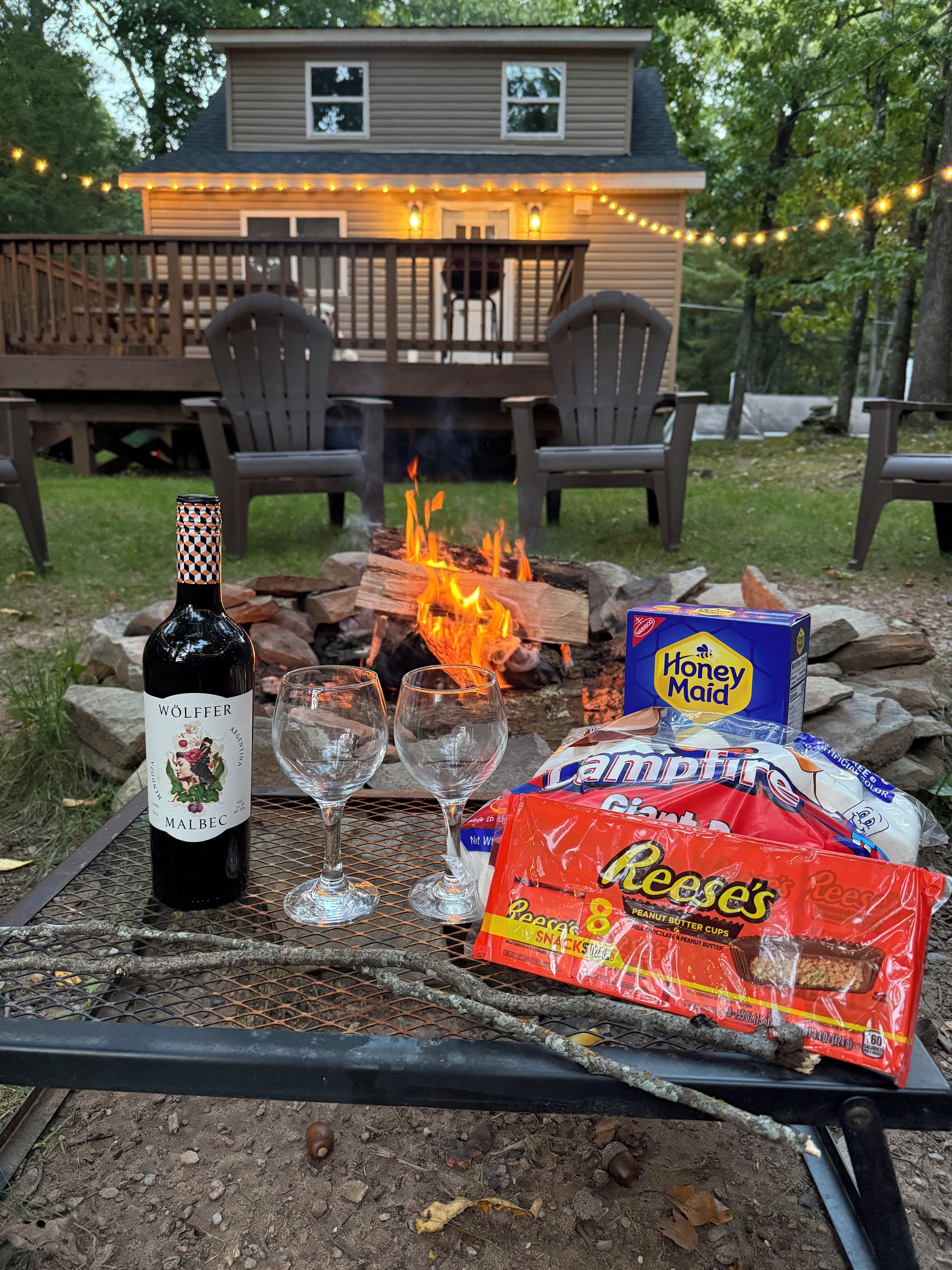 A bottle of wine, two glasses, a fire pit with logs, and snacks on a table outdoors.