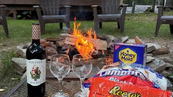 A bottle of wine, two glasses, a fire pit with logs, and snacks on a table outdoors.