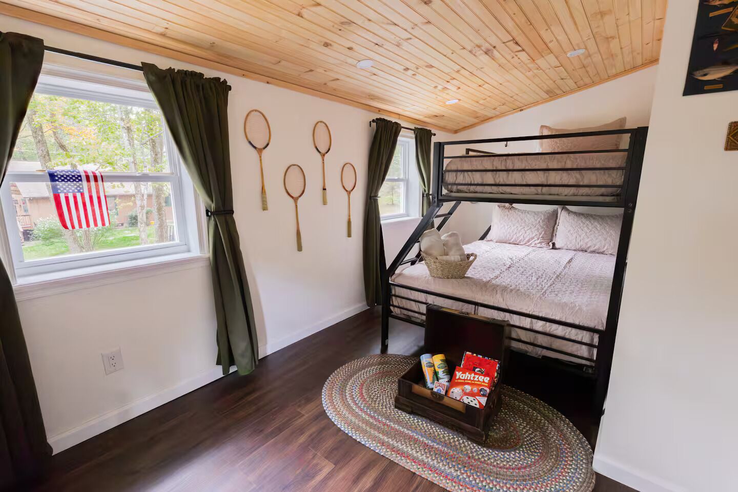 A bunk bed room with a wooden ceiling, a window with curtains, and a wall with badminton rackets.