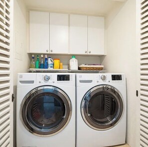 A laundry room with two front-loading washing machines and a shelf above containing various cleaning supplies. - Spacious 3-bedroom condo in a resort like community in Indian Wells. (Indian Wells)