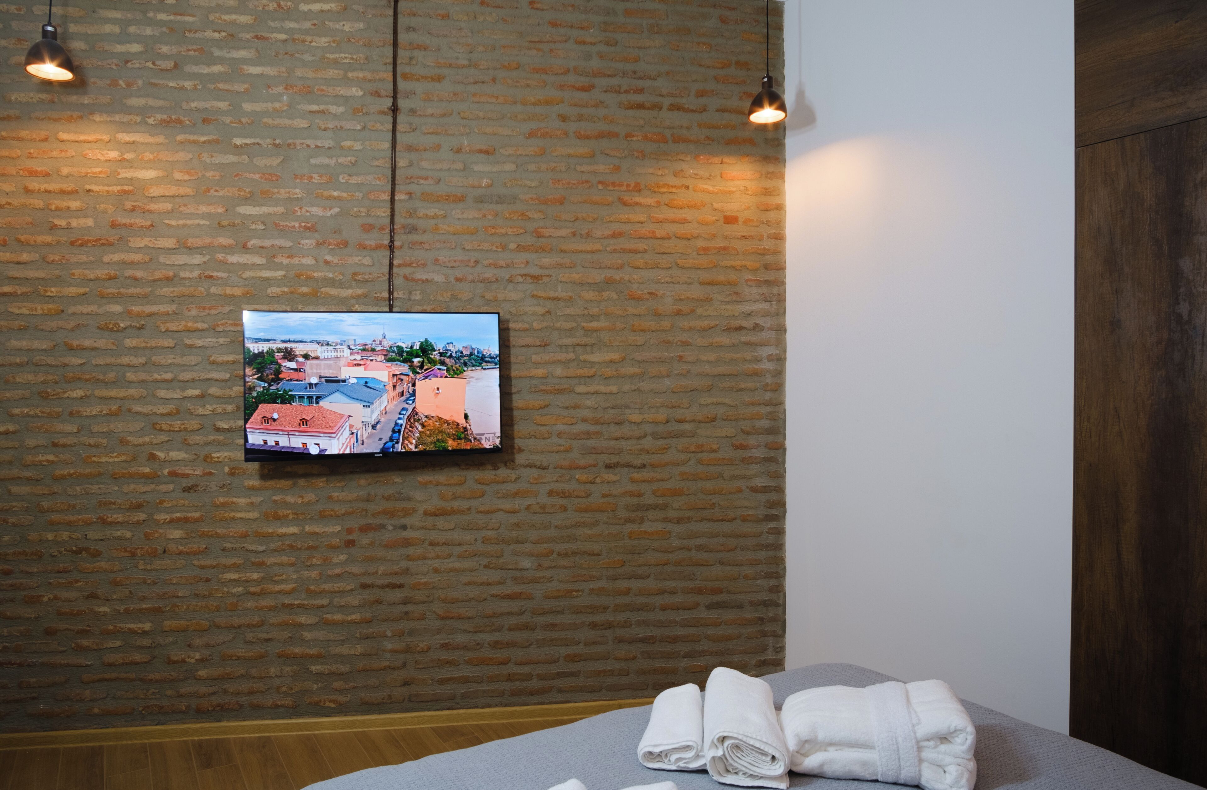 Une chambre d’hôtel avec un mur en briques, un téléviseur à écran plat fixé au mur, un lit recouvert d’une couverture grise et deux lampes suspendues.