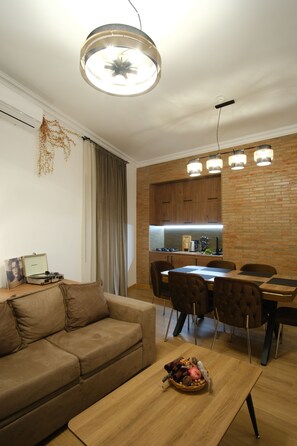 A modern living room with a sofa, a dining table with chairs, a kitchen area with a brick wall, and a ceiling light fixture.