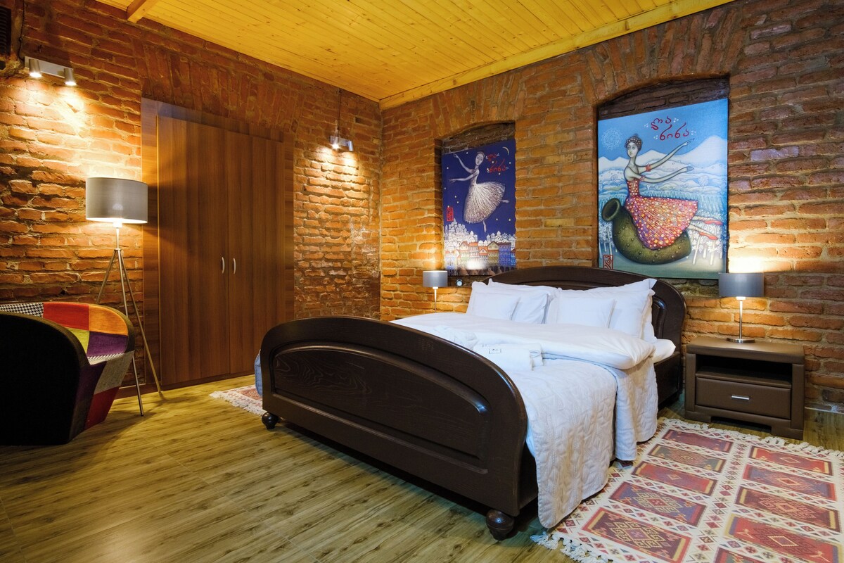 A bedroom with a wooden bed, a nightstand, a lamp, a chair, and a wall with a painting.