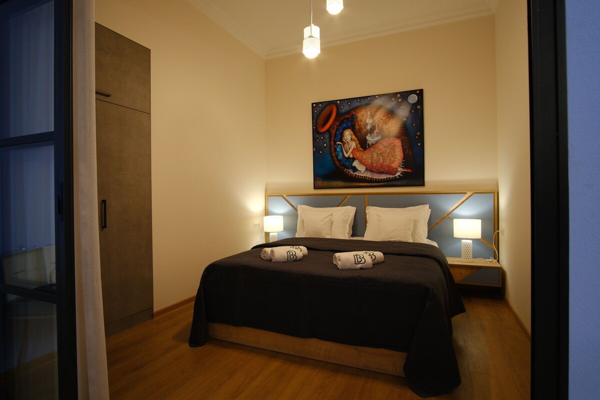 A hotel room with a large bed, two bedside lamps, a painting above the bed, and a wooden floor.