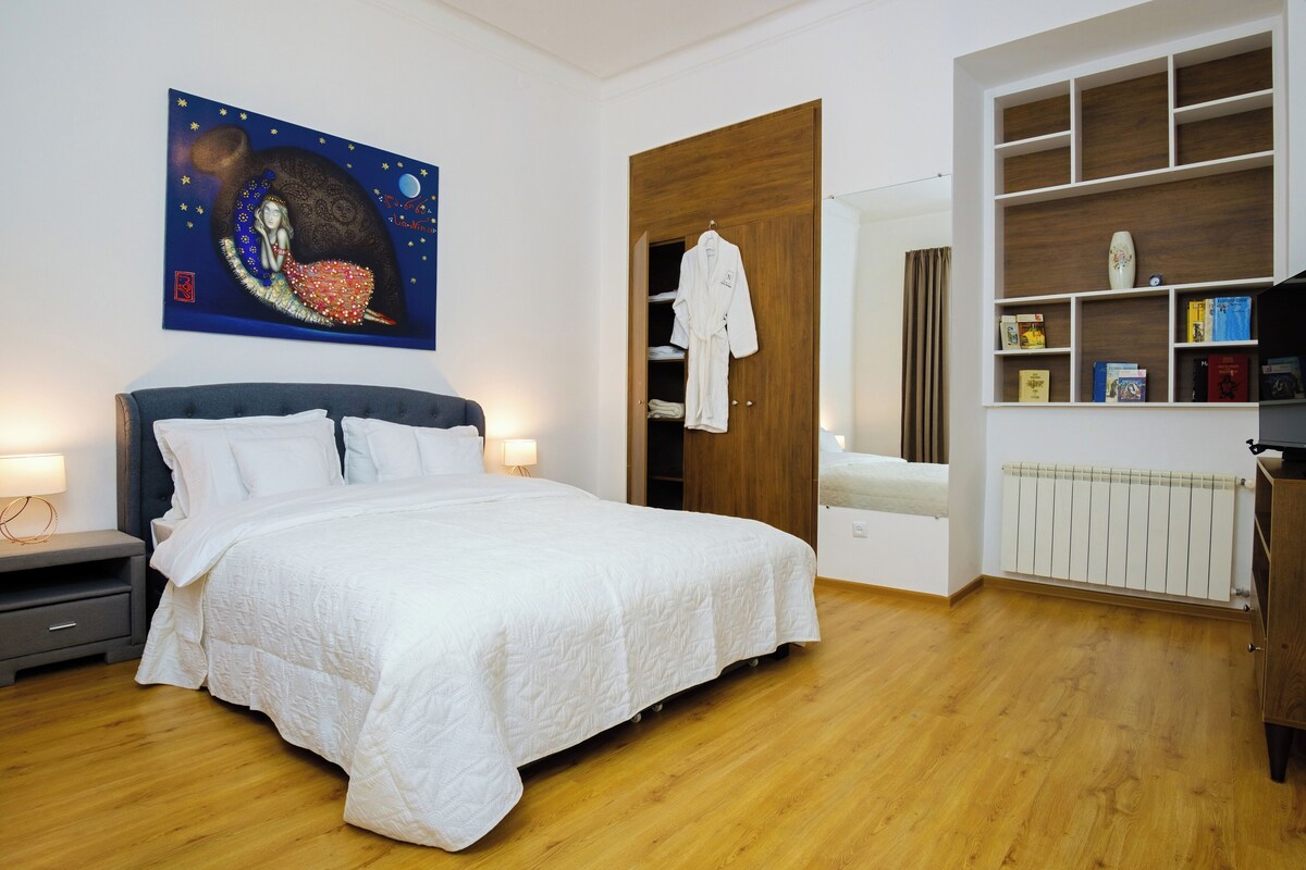 A bedroom with a large bed, a wardrobe, a bookshelf, and a painting on the wall.