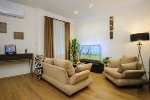 Una sala de estar con dos sofás beige, un televisor de pantalla plana, una planta y una lámpara de pared.