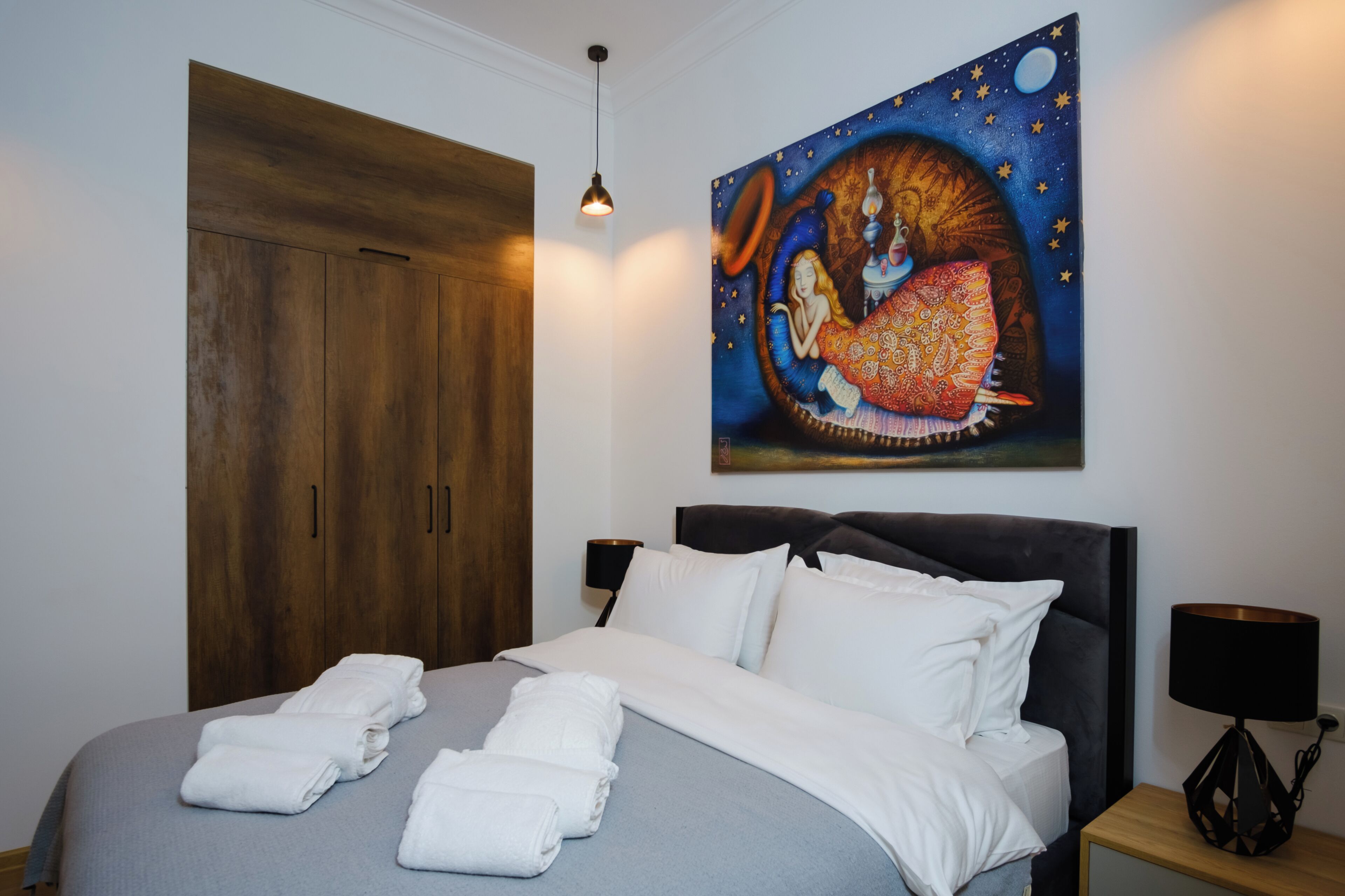 Quarto de hotel com uma cama grande, um guarda-roupa, um quadro de uma mulher com um vestido colorido e uma mesa de cabeceira com abajur.