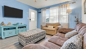A living room with a sofa, ottoman, and a television on the wall. - Beach Bungalow Near Ormond Beach and the Atlantic (Ormond Beach)
