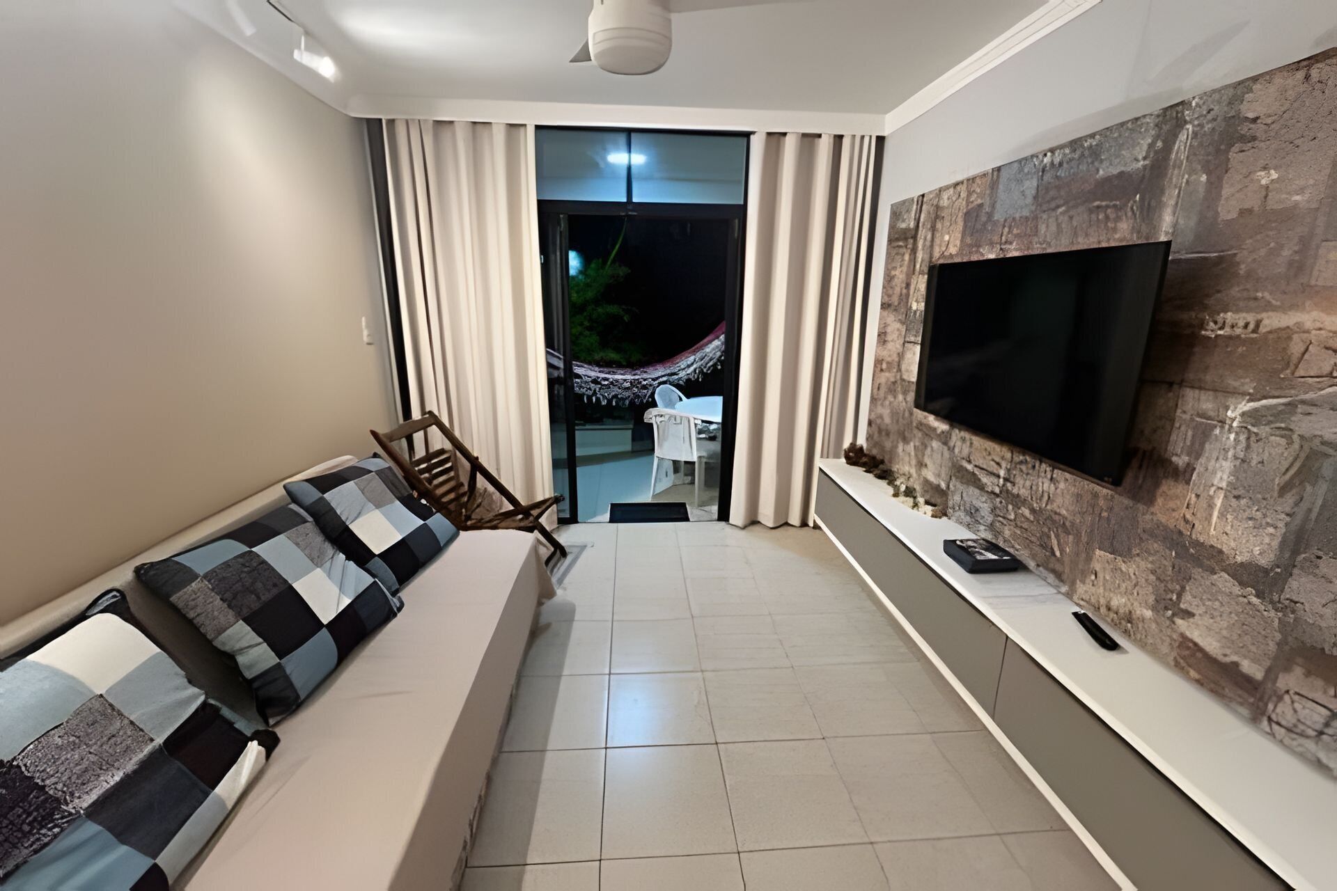 A modern living room with a sofa, a flat-screen TV mounted on a textured wall, and a view of an outdoor area with a hammock.