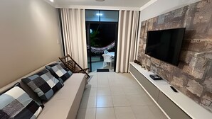 Un salón moderno con un sofá, un televisor de pantalla plana montado en una pared con textura, y vistas a un espacio exterior con una hamaca.