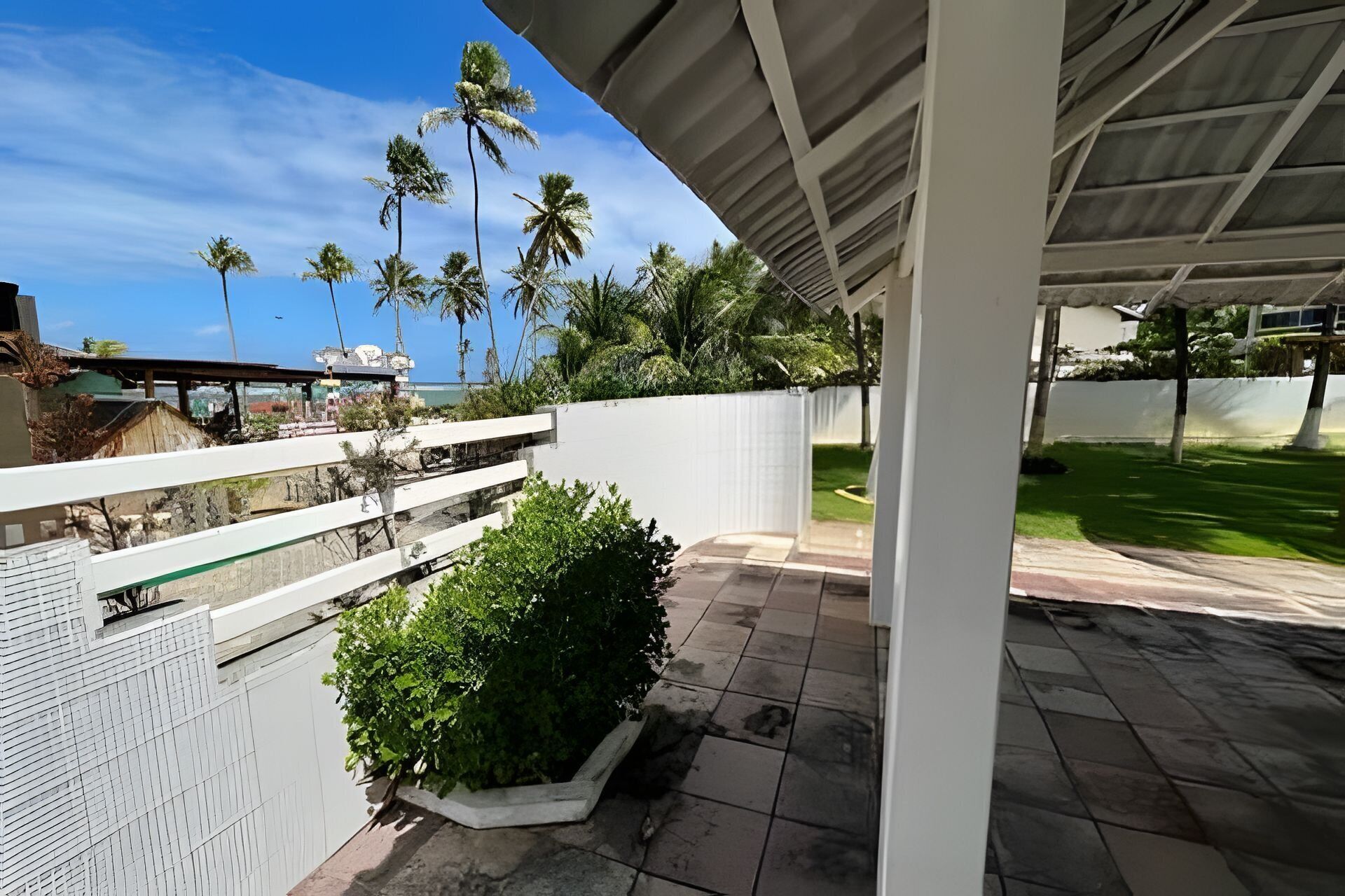 A covered outdoor area with a white fence, a paved patio, and a view of palm trees and a building in the background.