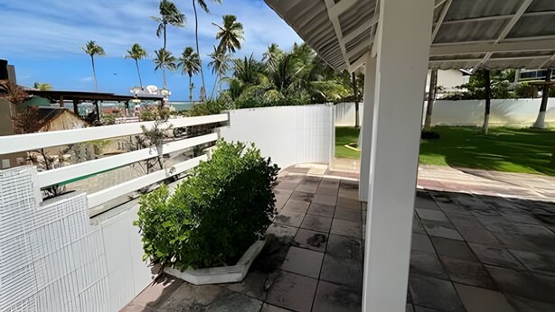 Un espacio al aire libre techado con una cerca blanca, un patio empedrado y vistas a palmeras y un edificio al fondo.