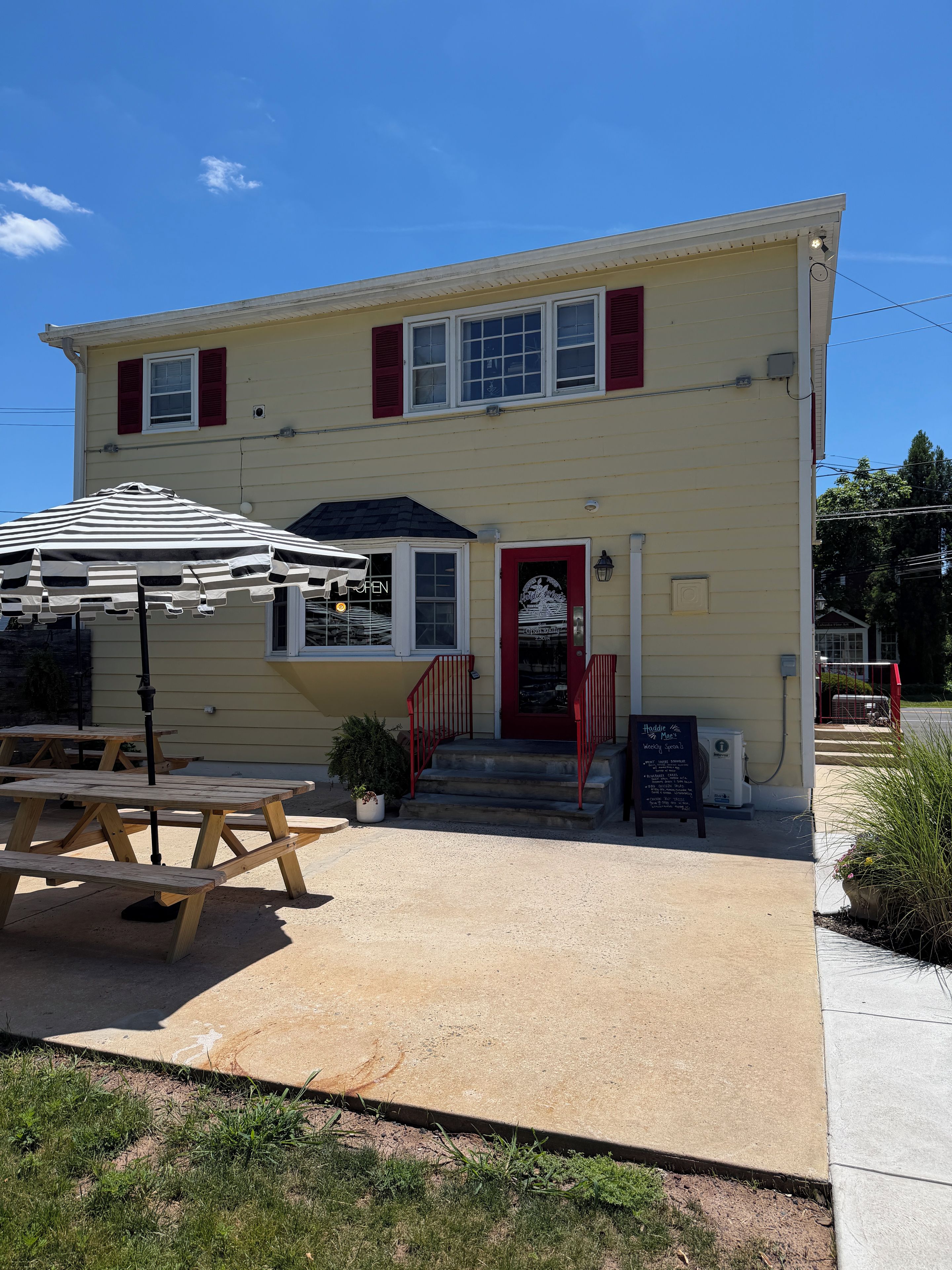 A two-story building with a red door, a small outdoor seating area with tables and an umbrella, and a signboard.