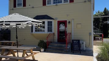 A two-story building with a red door, a small outdoor seating area with tables and an umbrella, and a signboard.