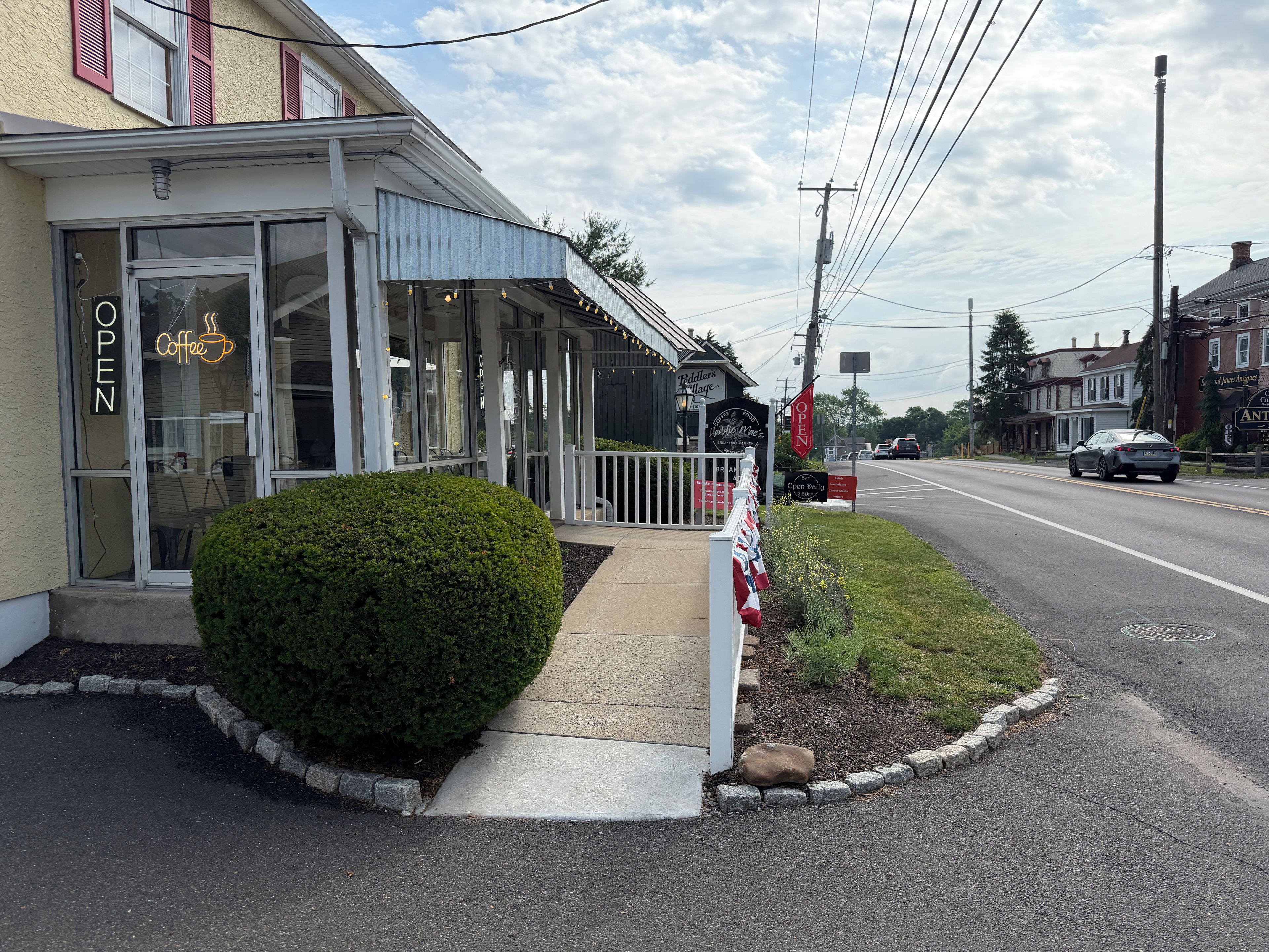 A storefront with a neon sign, a sidewalk, a small hedge, and a street with vehicles.