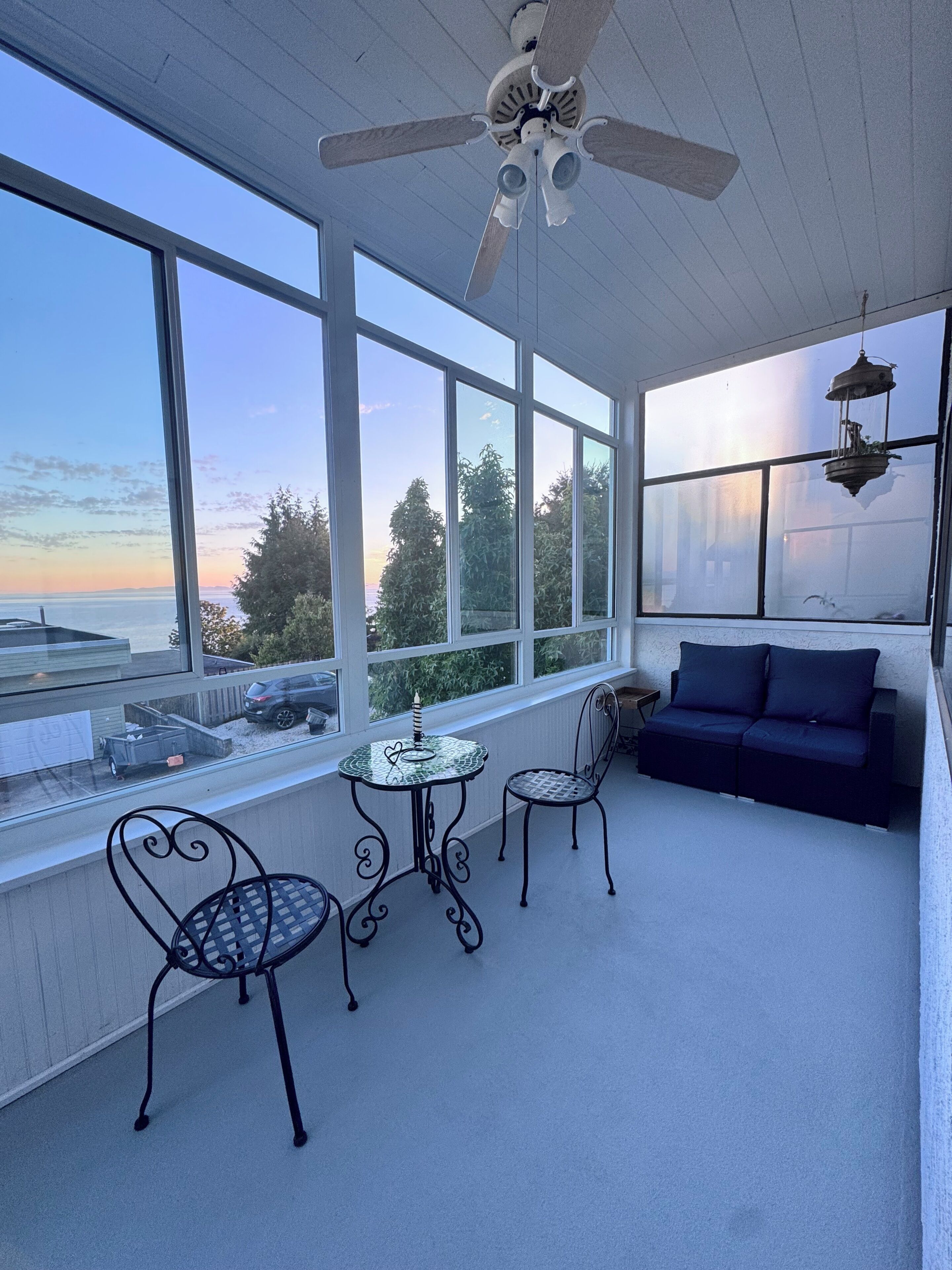 Una habitación iluminada por el sol con ventilador de techo, ventanas amplias, una mesa con sillas y un sofá.