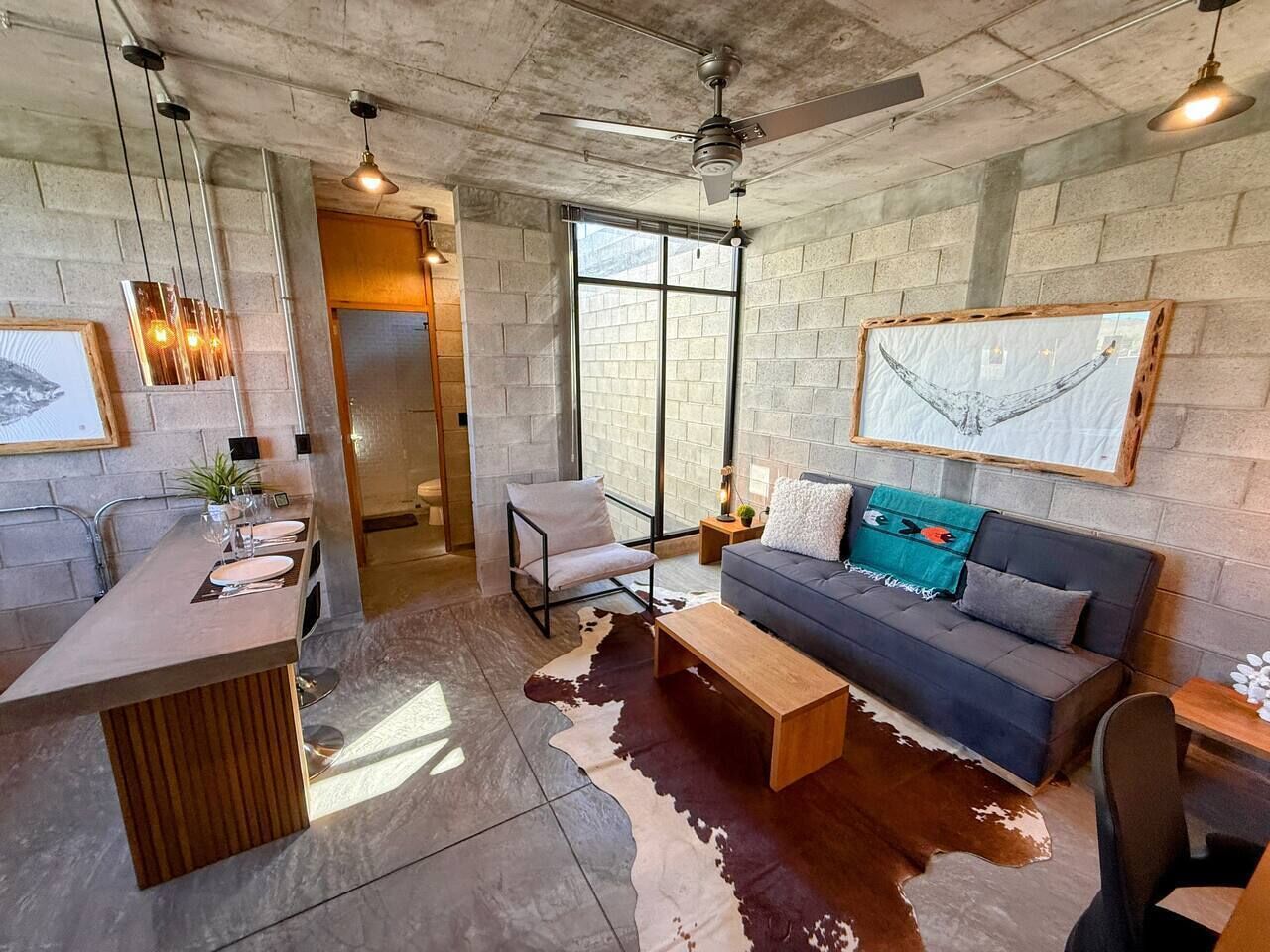 Un interior moderno con cocina, comedor y sala de estar, destacando un tapete de cuero de vaca, un sofá con cojines y una obra de arte colgante.