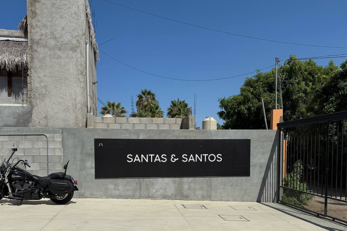 Ein Schild mit der Aufschrift 'SANTAS & SANTOS' an einer grauen Wand, davor ein abgestelltes Motorrad und ein verschlossenes Tor.
