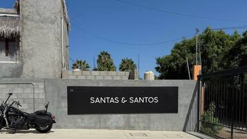 Ein Schild mit der Aufschrift 'SANTAS & SANTOS' an einer grauen Wand, davor ein abgestelltes Motorrad und ein verschlossenes Tor.
