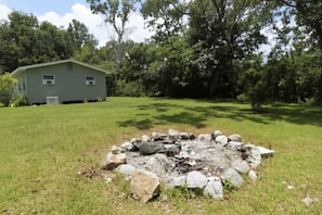 A grassy area with a stone fire pit surrounded by rocks, a small shed, and dense trees in the background.