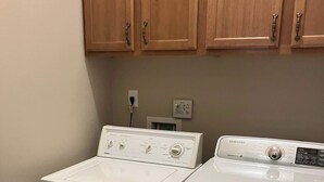 Un área de lavandería con dos lavadoras blancas, un cesto de ropa azul, armarios de madera arriba y un enchufe de pared.