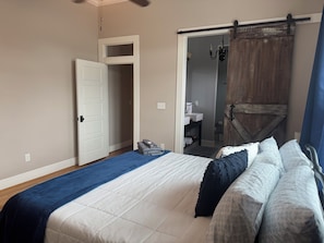 Quarto com cama, guarda-roupa, banheiro visível por uma porta aberta e ventoinha no teto.