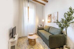 Een woonkamer met een bank, een klein rond tafeltje, een tv op een standaard, een potplant en kunst aan de muur.