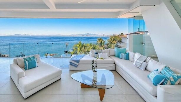 Een moderne woonkamer met een grote glazen tafel, witte banken en uitzicht op zee.