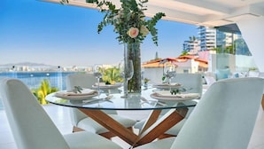 Een ronde glazen tafel met witte stoelen, een vaas met bloemen en een helder uitzicht op de zee en gebouwen op de achtergrond.