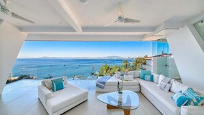 Een moderne woonkamer met een groot raam met uitzicht op de zee, ingericht met witte banken en een glazen salontafel.