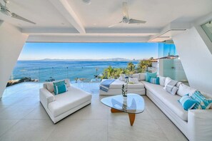 A modern living room with a large glass window overlooking the sea, furnished with white sofas and a glass coffee table. - Luxurious 2BR Condo - Amazing Ocean View - Pool (Puerto Vallarta)