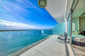 A balcony with a glass railing overlooking a body of water, a bed, and a chair. - Beachview Luxury 3 BR Condo - Pools - Beach (Puerto Vallarta)