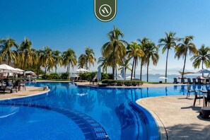 A swimming pool with lounge chairs and umbrellas, surrounded by palm trees and a view of the ocean. - B Nayar 3BR Sanctuary: Private Balcony, Full Kitchen & Steps to La Cruz Marina (La Cruz de Huanacaxtle)