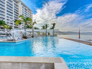 Ein Hotelpool mit klarem, blauem Wasser, umgeben von Liegestühlen und Sonnenschirmen, mit Blick auf das Meer und einem Gebäude im Hintergrund.
