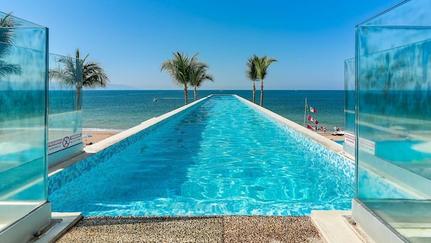 Ein langer Swimmingpool mit klarem, blauem Wasser, umgeben von Palmen und Blick auf den Ozean.