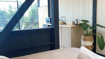 Ein Schlafzimmer mit einem Bett, Kissen, einem Fenster, einer Küchenzeile und einer Topfpflanze.