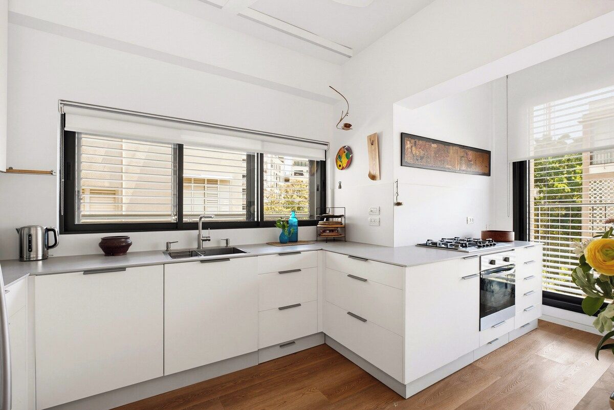 Une cuisine moderne avec des meubles blancs, un four encastré et une grande fenêtre équipée de stores.