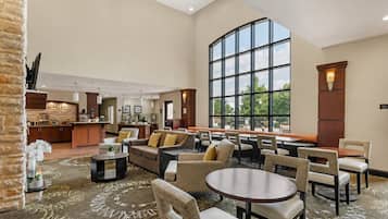 A spacious hotel lobby with a dining area, a kitchen in the background, and large windows offering a view.
