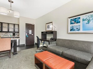 A modern living room with a sofa, a coffee table, and a desk with a chair. There is a kitchen area with cabinets and a countertop.
