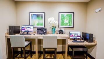 A hotel room with a desk, two computers, a printer, a vase with flowers, and framed pictures on the wall.