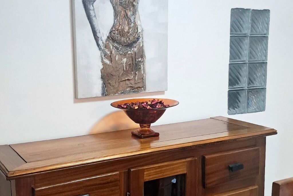 A wooden dresser with a glass bowl on top, a painting on the wall, and a textured wall panel.