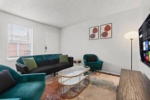 A living room with a black sofa, teal armchairs, a glass coffee table, and a wall-mounted TV. - Chic & Cozy Zilker Park 2BR | Walkable & Stylish (Austin)