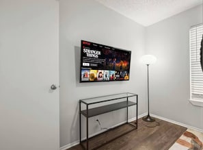 Quarto com televisão de tela plana na parede exibindo uma série da Netflix, uma mesinha de console de vidro, abajur e janela com persianas.