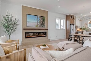 A modern living room with a fireplace, a television mounted on the wall, and a dining area with a wooden table. - Biển Kay – Luxury Beach House w/ Pool + Golf Cart (Holden Beach)