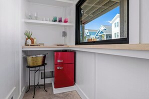 A compact kitchenette with a red refrigerator, a bar stool, and a window with a view of buildings. - Biển Kay – Luxury Beach House w/ Pool + Golf Cart (Holden Beach)