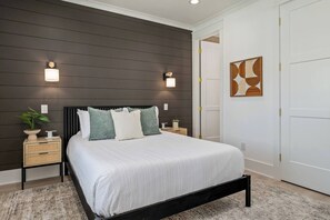A bedroom with a dark wood panel wall, a white bed with multiple pillows, bedside tables with lamps, and a door with a geometric artwork. - Biển Kay – Luxury Beach House w/ Pool + Golf Cart (Holden Beach)