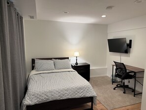 A bedroom with a bed, a bedside table with a lamp, a TV mounted on the wall, and a desk with a chair.